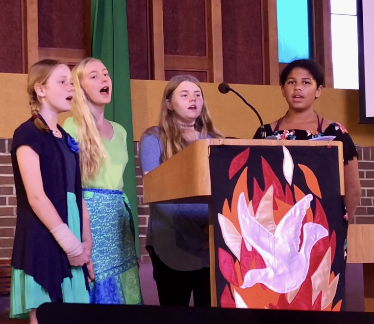 Four girls singing in front of a pulpit at a Sunday church service.